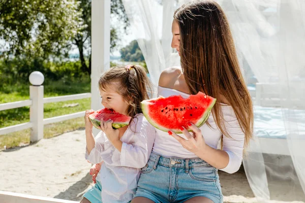 Plajda karpuz yiyen anne ile birlikte beyaz elbiseli mutlu neşeli küçük kız.