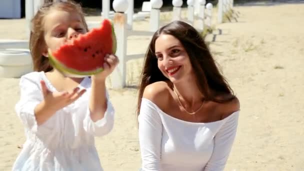 ビーチでスイカを食べる楽しみを持っているママと一緒に白いドレスで幸せな陽気な小さな女の子 — ストック動画
