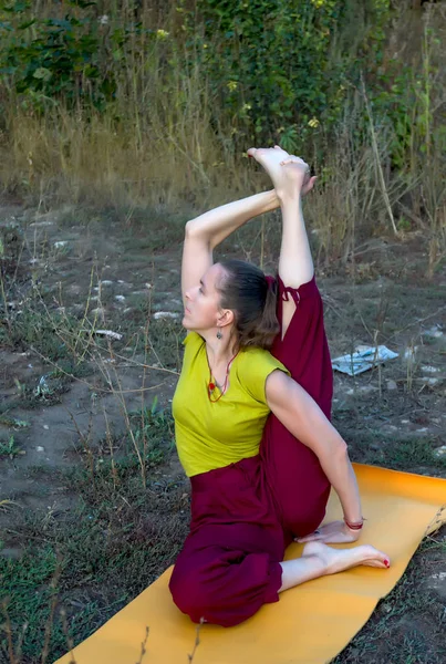 A woman practicing yoga in nature and sitting in twine lifting one leg up over her head