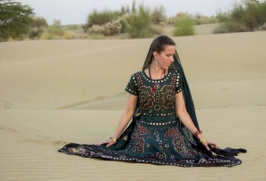 Ulusal Hint kostümlü genç bir kadın, Thar çölündeki kum tepelerinin arasında düşünceli bir şekilde kuma oturur. Rajasthan, Hindistan.