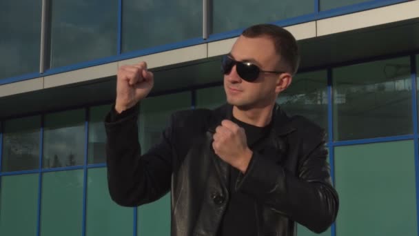 Молодий чоловік у шкіряній куртці та сонцезахисних окулярах стоїть у позі боксера на відкритому повітрі — стокове відео