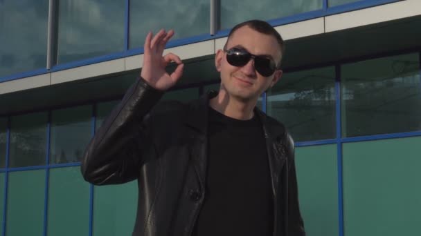 Jeune homme en veste en cuir et lunettes de soleil debout en plein air et montrant ok — Video