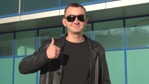 Молодой человек в кожаной куртке и солнцезащитных очках стоит на улице и показывает большой палец вверх — стоковое видео