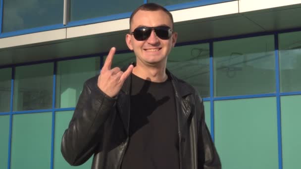 Jeune homme en veste en cuir et lunettes de soleil debout en plein air et montre le signe des cornes — Video