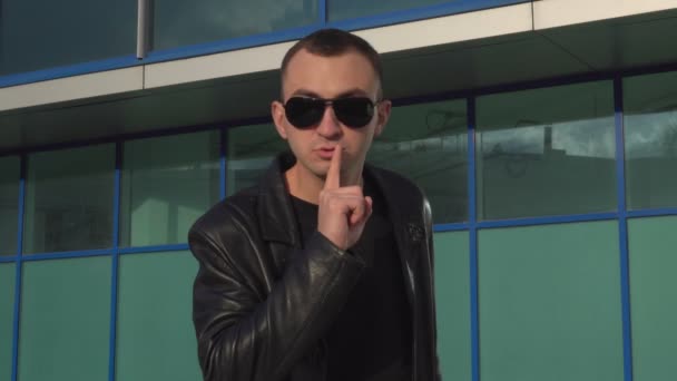Молодой человек в кожаной куртке и солнцезащитных очках стоит на улице и делает жест молчания — стоковое видео