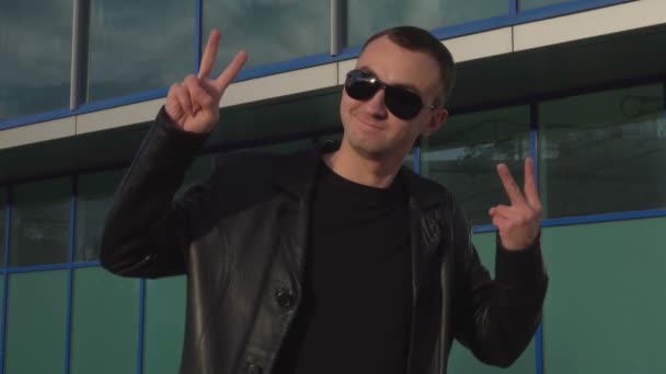 Hombre joven en chaqueta de cuero y gafas de sol de pie al aire libre y haciendo señal de paz — Vídeos de Stock