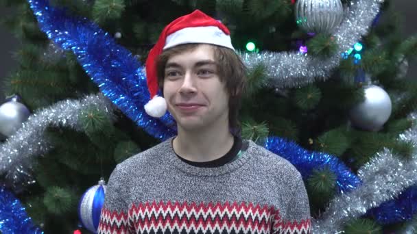 Retrato del joven sonriente en Santa Hat mirando a la cámara en el fondo del árbol de Navidad . — Vídeo de stock