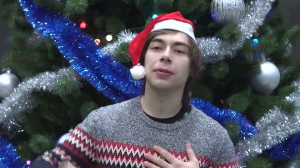 Bonito jovem vestindo chapéu de Papai Noel e falando sobre algo no fundo da árvore de Natal . — Vídeo de Stock