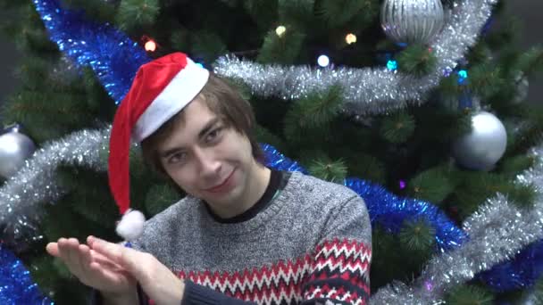 Knappe jonge Man dragen kerstmuts en Is klappen op kerstboom achtergrond. — Stockvideo