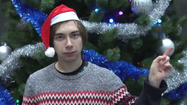 Hombre joven guapo con sombrero de Santa y dedo ondeante para rechazar en el fondo del árbol de Navidad . — Vídeos de Stock