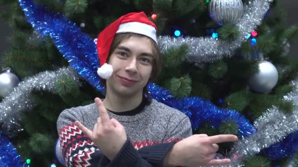 Hombre joven guapo con sombrero de Santa y haciendo gestos de roca en el fondo del árbol de Navidad . — Vídeo de stock