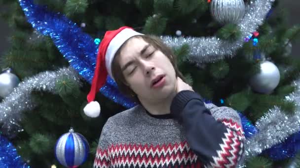 Porträtt av ung Man i Santa Hat Witn smärta i halsen på julgran bakgrund — Stockvideo