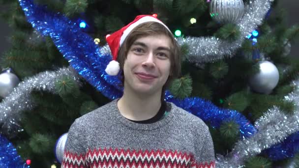 Retrato del joven sonriente en Santa Hat mirando a la cámara en el fondo del árbol de Navidad — Vídeo de stock