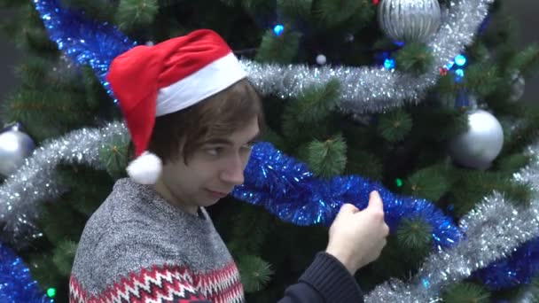 Hombre joven guapo con sombrero de Santa e invitando a alguien a venir en el fondo del árbol de Navidad . — Vídeo de stock