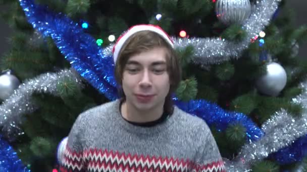 Guapo joven está usando Santa Sombrero y está bailando en el fondo del árbol de Navidad . — Vídeo de stock