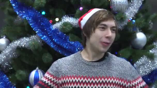 Guapo joven con sombrero de Santa y gritando sorprendido agradable choque en el fondo del árbol de Navidad . — Vídeo de stock