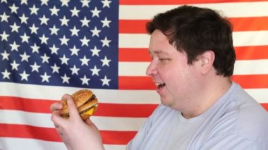 Amerikan bayrağı geçmişi olan bir parça hamburger tutan şişman adam. Büyük adam fast food istiyor.