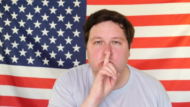 Bir Amerikan bayrağının arka planında sessizlik jesti gösteren genç bir adam. — Stok video
