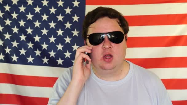 Hombre gordo joven en gafas de sol hablando por teléfono en el fondo de una bandera de EE.UU.. — Vídeo de stock