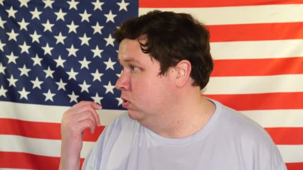 Zijaanzicht van jonge dikke man die iemand uitnodigt op de achtergrond van een Amerikaanse vlag — Stockvideo