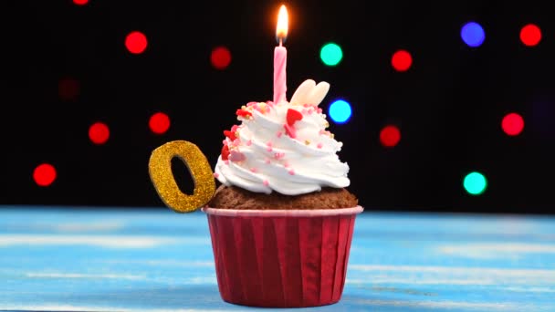 Delicious birthday cupcake with burning candle and number 0 on multicolored blurred lights background — Stock Video