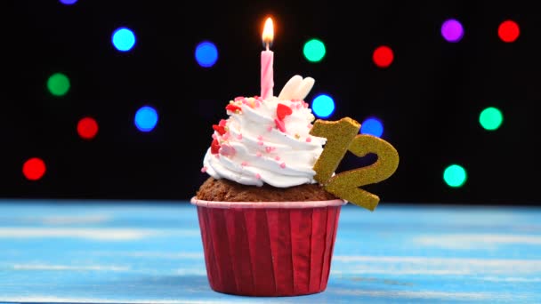 Delicious birthday cupcake with burning candle and number 12 on multicolored blurred lights background — Stock Video