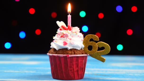 Вкусный праздничный кекс с горящей свечой и цифра 62 на многоцветном фоне размытых огней — стоковое видео