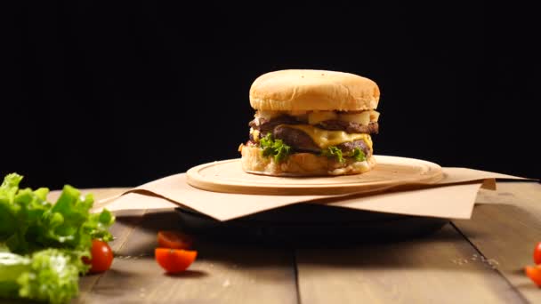 Hamburger frais juteux au fromage, sauce et légumes tournant sur la planche de bois sur fond noir — Video