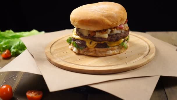 Grand burger juteux au fromage, sauce et légumes tournant sur la planche de bois sur fond noir — Video