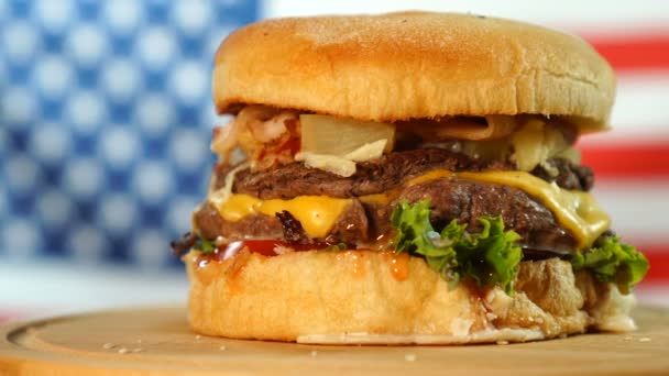 Βοδινό ζουμερό burger με τυρί, σάλτσα και λαχανικά που περιστρέφονται σε μια ξύλινη σανίδα μπροστά από την αμερικανική σημαία — Αρχείο Βίντεο