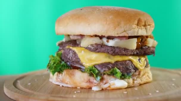 Velký burger se sýrem, omáčkou a zeleninou se rychle otáčí na dřevěné desce před zeleným pozadím. — Stock video