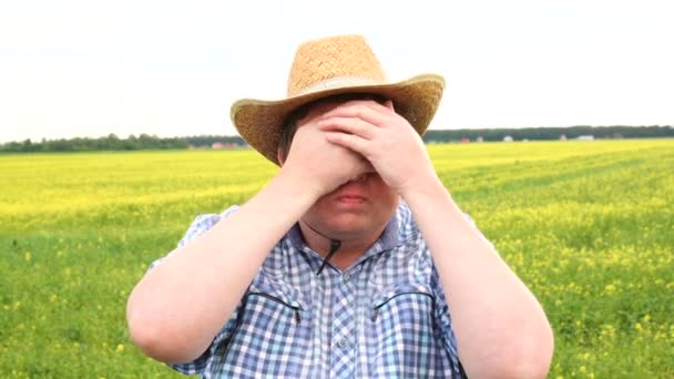 Joven frustrado con camisa y sombrero cubriendo los ojos cerrados mientras se paraba contra el campo en un día soleado — Vídeo de stock