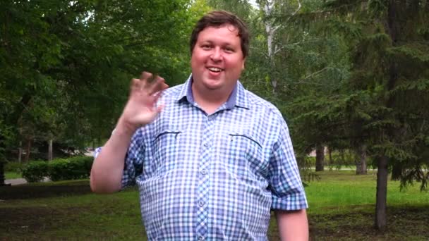 Hombre gordo con camisa a cuadros saludando con la mano con expresión feliz en un parque — Vídeos de Stock