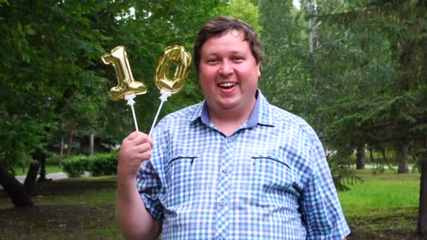 Grande homem segurando dois balões dourados fazendo o número 10 ao ar livre. Festa de comemoração do décimo aniversário — Vídeo de Stock