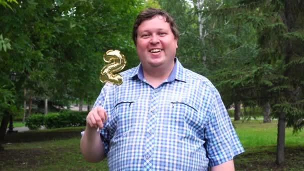 Velký muž, který drží zlatý balónek, dělá 2 číslo venku. Oslava narozenin 2. — Stock video