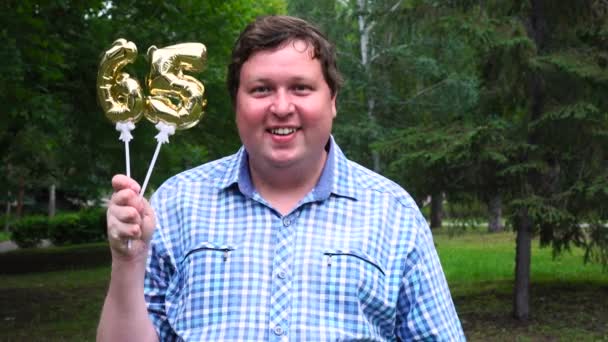 Big man innehar gyllene ballonger gör 65 nummer utomhus. 65-års jubileum fest — Stockvideo
