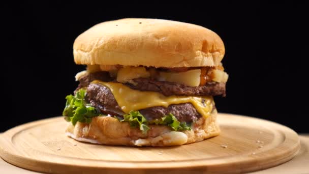 Burger, with a beef, cheese, and vegetables rotates on a wooden Board. On black background — Stock Video