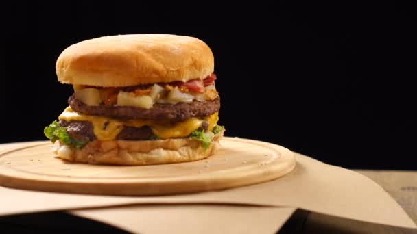 La hamburguesa, con una carne de res, queso y verduras gira sobre una mesa de madera. Sobre fondo negro — Vídeos de Stock