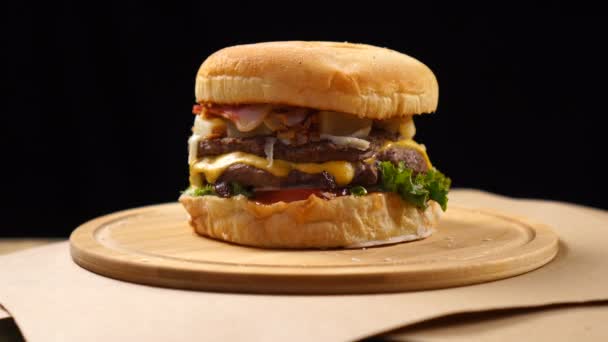 Hamburguesa de ternera americana a la parrilla con lechuga, queso, cebolla servida sobre trozos de papel marrón girando sobre un mostrador de madera . — Vídeos de Stock