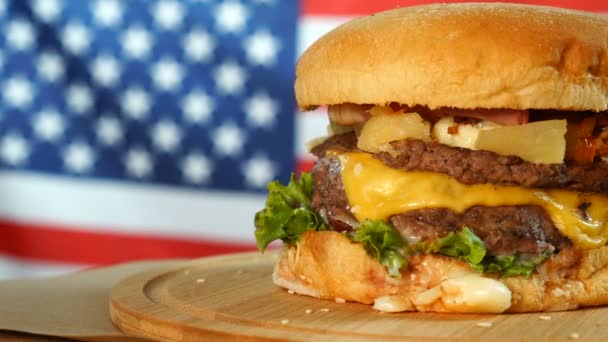 Hamburguesa de ternera americana a la parrilla con lechuga, queso, cebolla servida sobre trozos de papel marrón girando sobre un mostrador de madera . — Vídeos de Stock