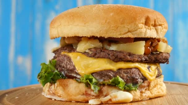 La hamburguesa, con una carne de res, queso y verduras gira sobre una mesa de madera. Sobre fondo de madera azul — Vídeo de stock