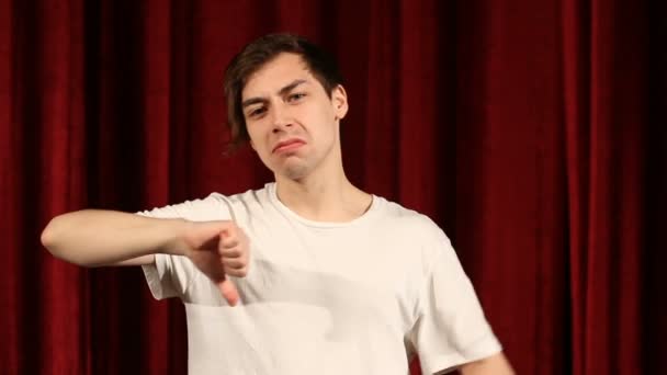 Молодой человек показывает большой палец вниз на красном фоне занавески — стоковое видео