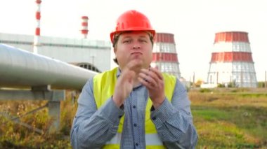İşçi, mühendis ya da elektrikçi doğrudan kameraya bakıyor. Elektrik santralinin önünde el çırpıyor.