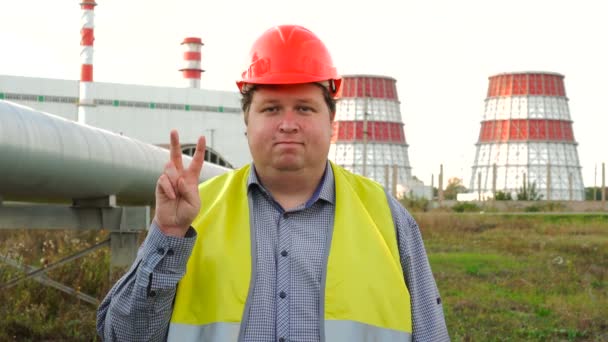 Pracownik, inżynier lub elektryk patrzący w kamerę pokazującą gest zwycięstwa stojący przed elektrownią — Wideo stockowe