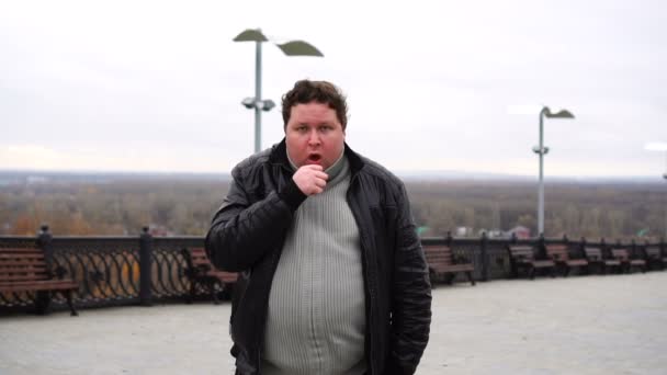 추운 가을 날씨에 야외에서 카메라와 기침을 하고 있는 뚱뚱 한 남자의 사진을 확대 해 보 세요.. — 비디오