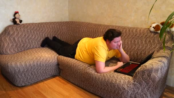 Gordo usando laptop deitado no sofá na sala de estar. Jovem navegando na internet, navegando na web, lendo notícias online — Vídeo de Stock