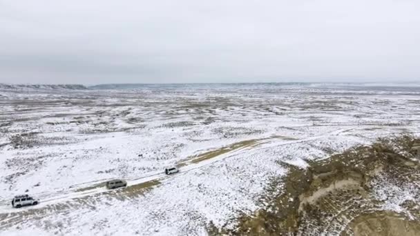 Três SUVs movendo-se ao longo da borda de um penhasco arenoso do deserto coberto de neve no inverno. Cazaquistão Ocidental, Península de Mangyshlak . — Vídeo de Stock