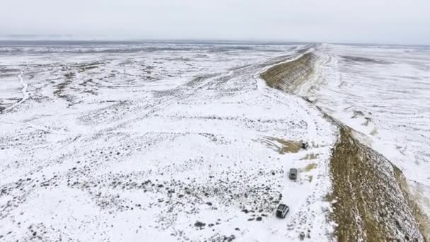 Três SUVs movendo-se ao longo da borda de um penhasco arenoso do deserto coberto de neve no inverno. Cazaquistão Ocidental, Península de Mangyshlak . — Vídeo de Stock
