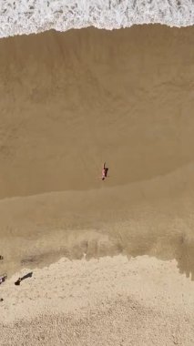 Güzel 4K dikey drone, okyanus dalgaları yavaşça kıyıya yaklaşırken sıcak kumda yatan hamile bir kadının görüntüsünü yakınlaştırıyor. Yukardan çekilen huzurlu bir annelik anı, sükunet, doğal güzellik ve zamansız uyum dolu..