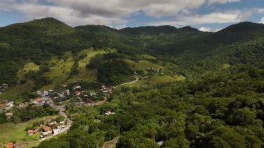 Hava aracı görüntüleri yemyeşil dağların üzerinde yavaşça uçuyor ve Brezilya 'nın Santa Catarina kentindeki kırsal bir köyde. Tepeler, ormanlar, evler ve bulutlu gökyüzü manzaralı.
